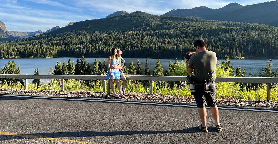 A photographer takes pictures of a couple standing by a scenic lake with forested mountains in the background in Bozeman, Montana.