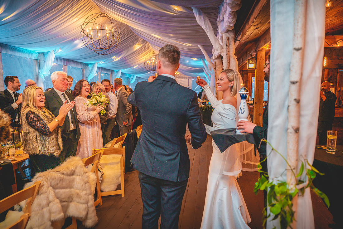 Bride and groom sharing a celebratory toast with guests at a beautifully decorated wedding venue in Bozeman, Montana. The venue features draped ceilings, warm lighting, and happy attendees applauding the couple.